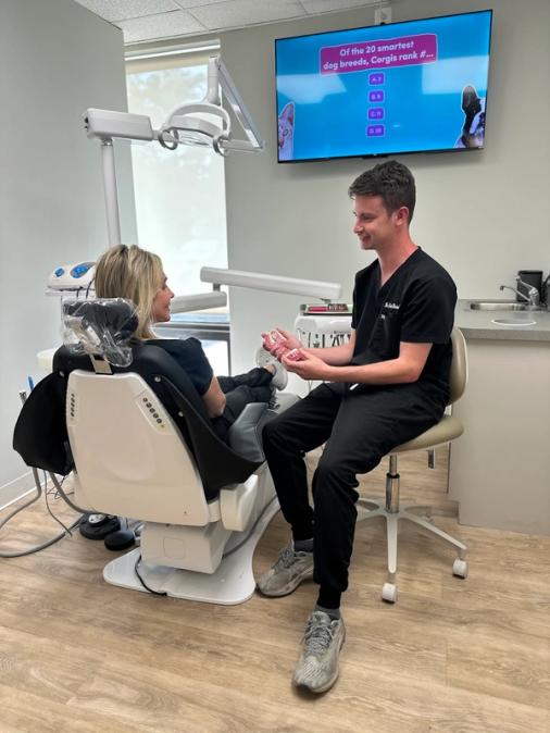 Dental professional discussing dental models with a patient in a dental chair, with a dog trivia game on a TV in the background.