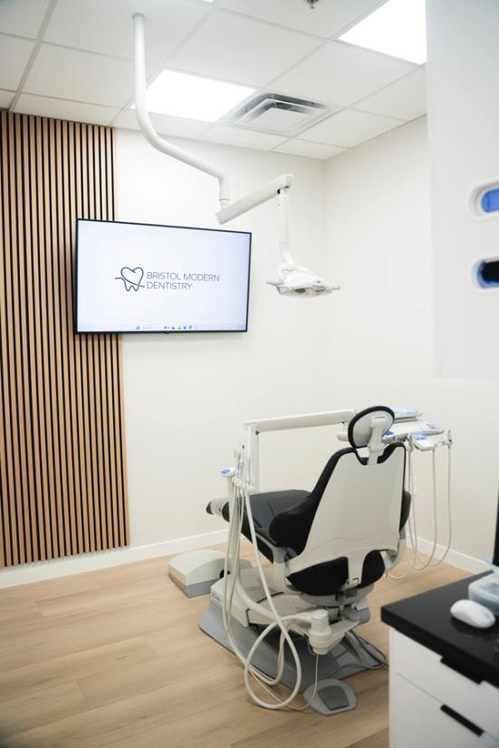 A modern dental office featuring a black and white dental chair, a TV displaying the "Bristol Modern Dentistry" logo, and a wood-slatted accent wall.