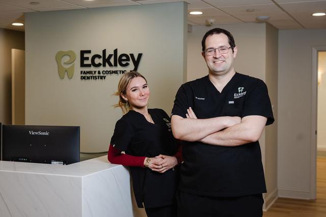 Two smiling dental professionals in scrubs stand in their office reception area, behind a counter with the "Eckley Family & Cosmetic Dentistry" sign visible on the wall.