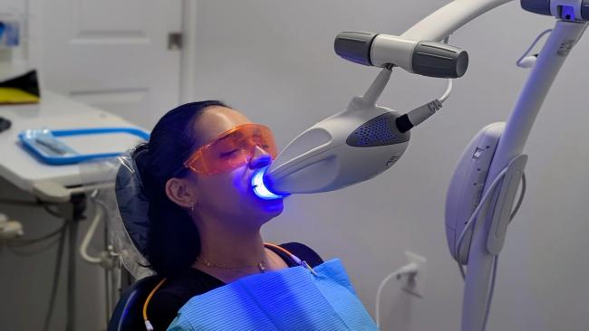 A woman wearing orange protective glasses undergoes blue light teeth whitening in a dental chair.