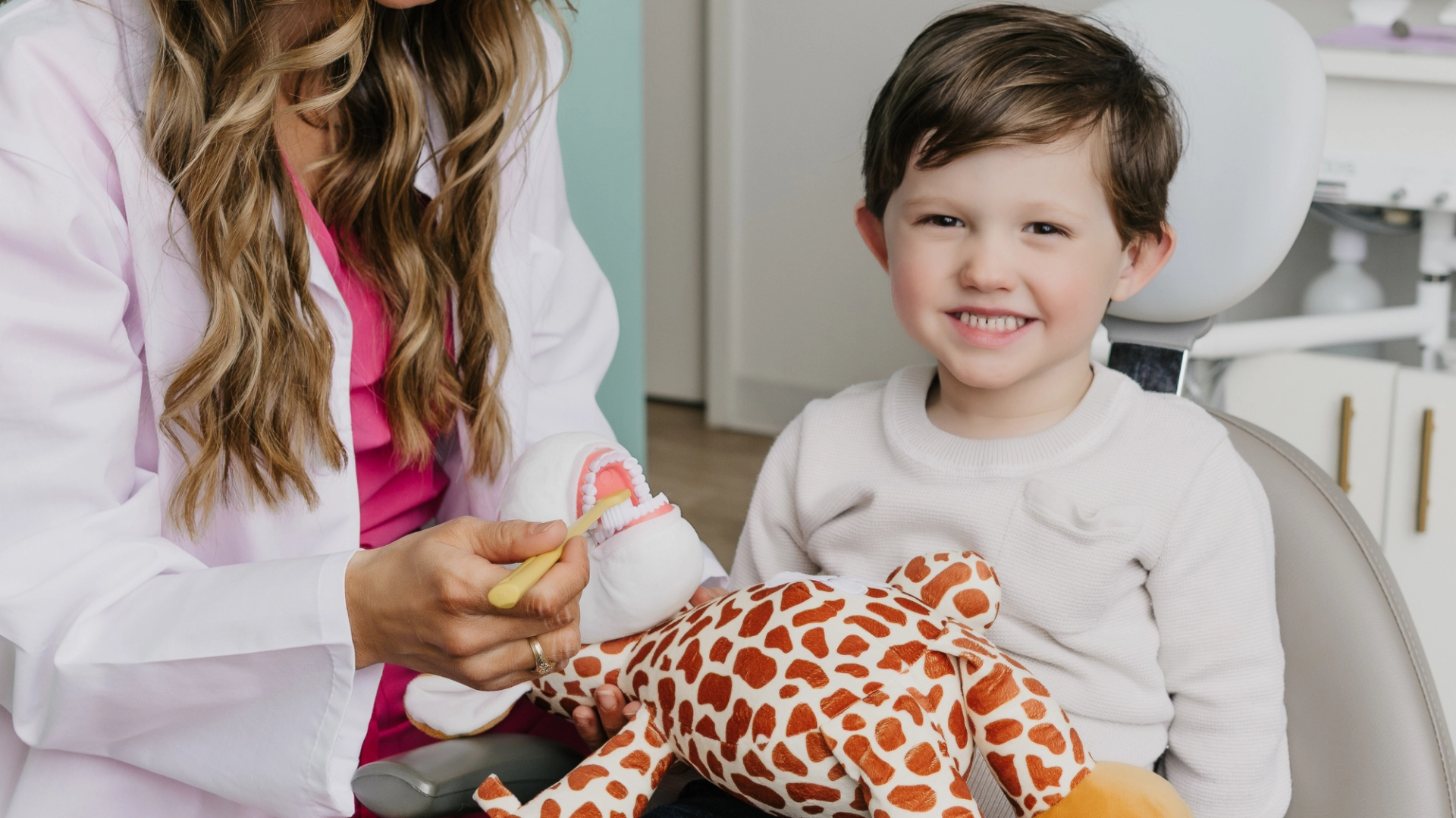 A dentist demonstrates teeth brushing on a model for a smiling child holding a giraffe toy.