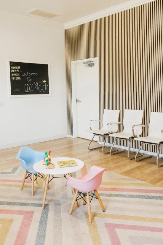 A modern waiting room with a blackboard, wood-slat accent wall, three white adult chairs, and a children's play area with a table and colorful chairs on a striped rug.