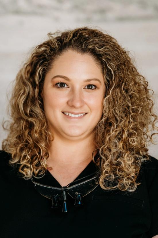 A smiling woman with curly light brown hair wears black scrubs with dental loupes around her neck.