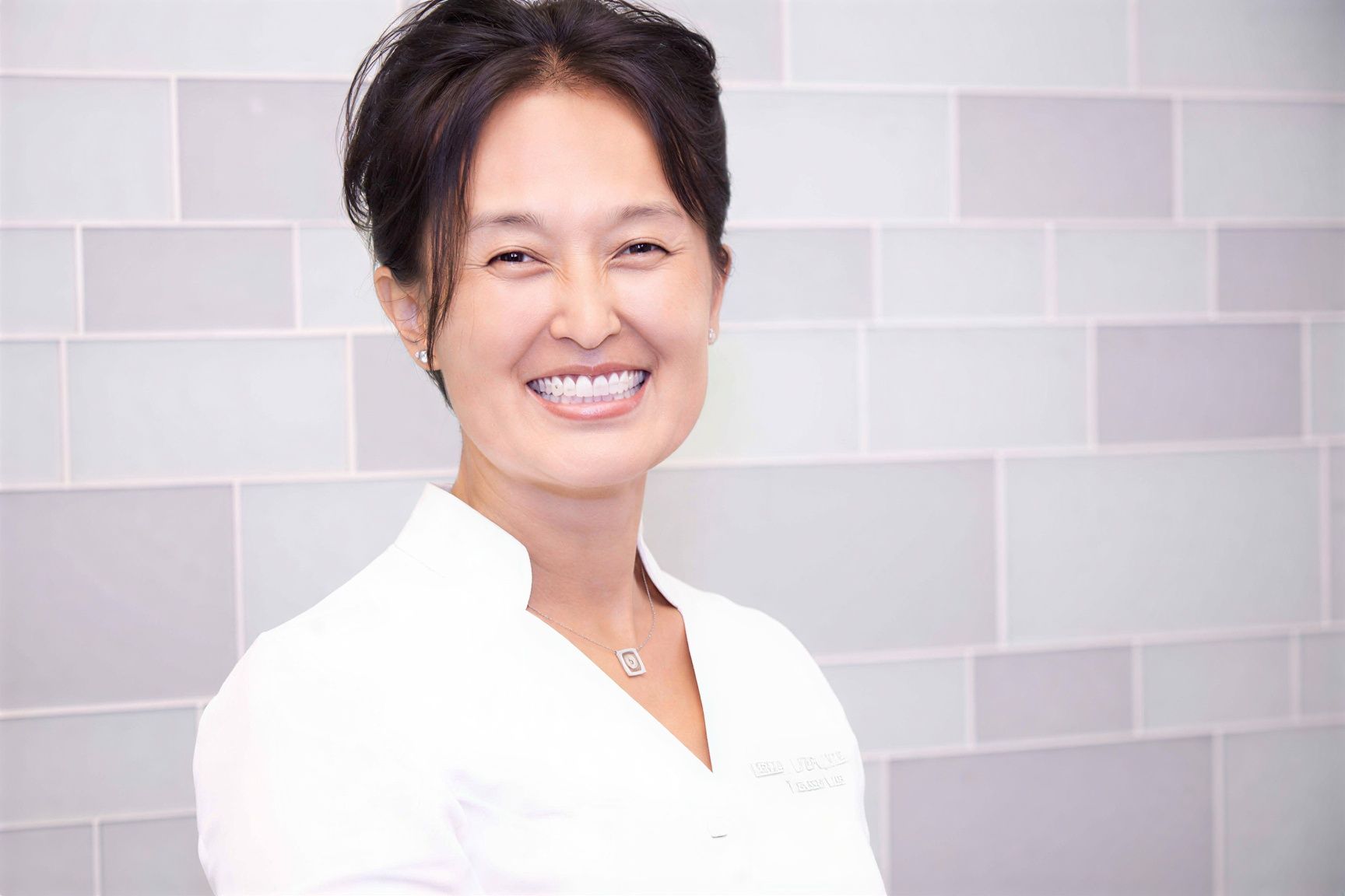 A smiling woman in a white uniform against a light gray tiled wall.