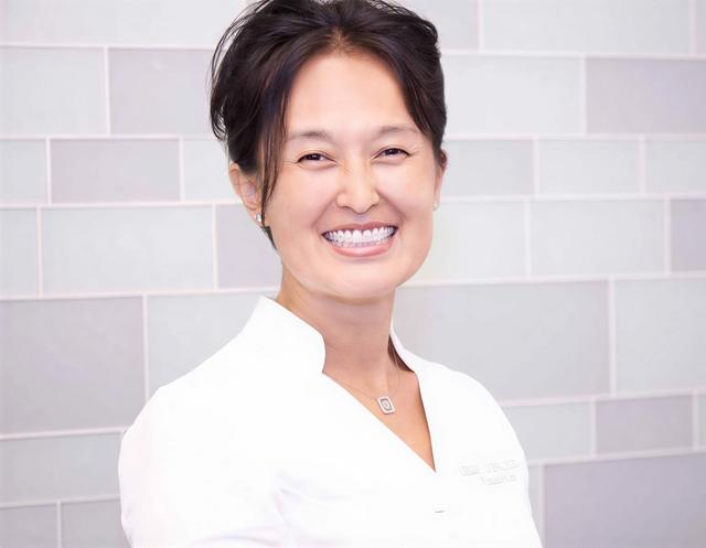 A smiling woman in a white uniform against a light gray tiled wall.