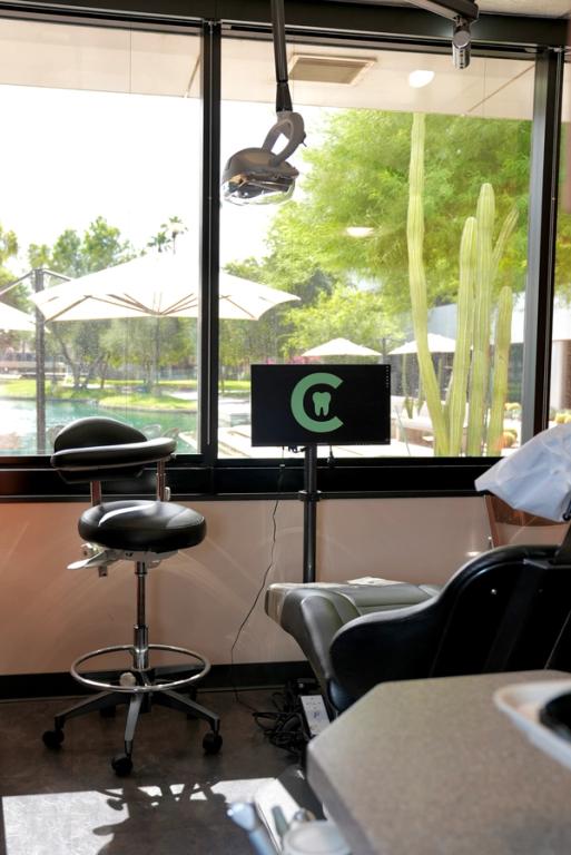 A dental office interior with a patient chair, a dentist's stool, a monitor displaying a tooth logo, and a large window overlooking a pond and desert plants.