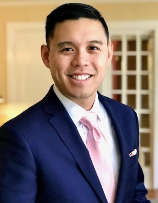 A smiling man in a dark blue suit with a pink tie.