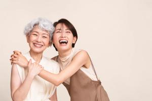 An older woman with grey hair smiles while a younger woman embraces her and laughs heartily.