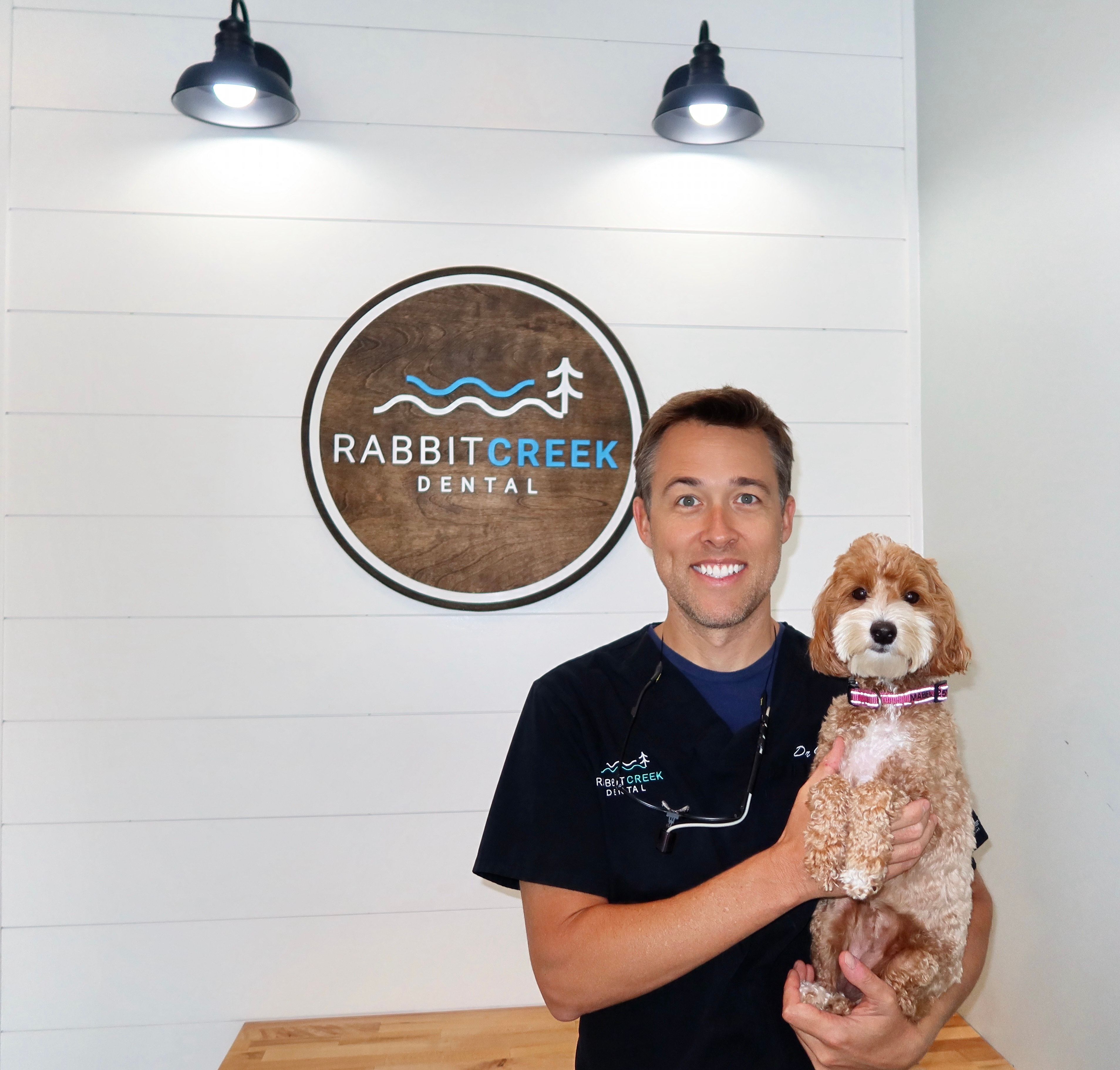 a man is holding a small dog in front of a rabbit creek dental sign .