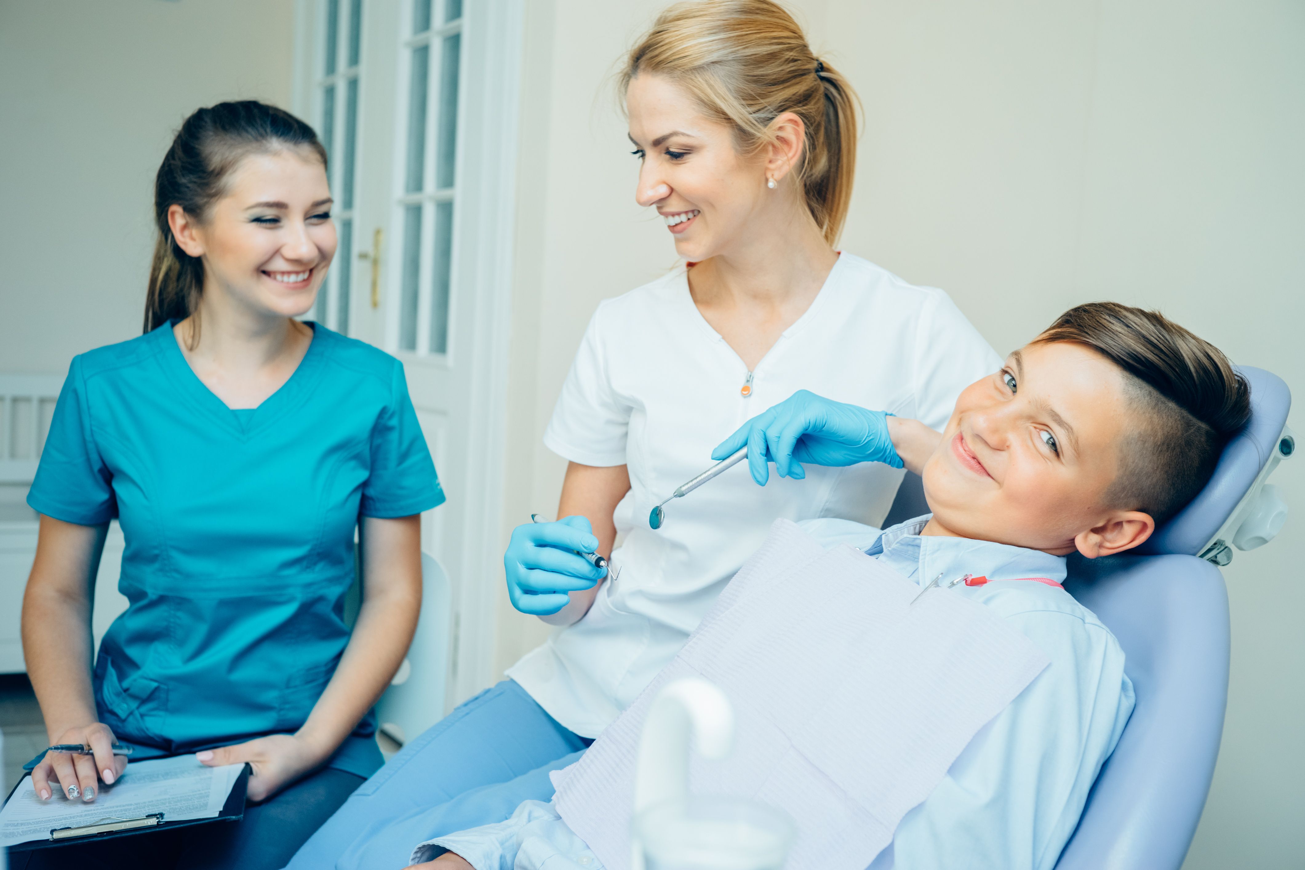 A smiling boy in a dental chair with a smiling dentist holding tools and an assistant.