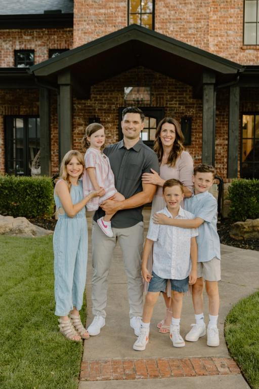 A happy family of six, with parents and four children, smiles for a photo in front of a modern brick house.
