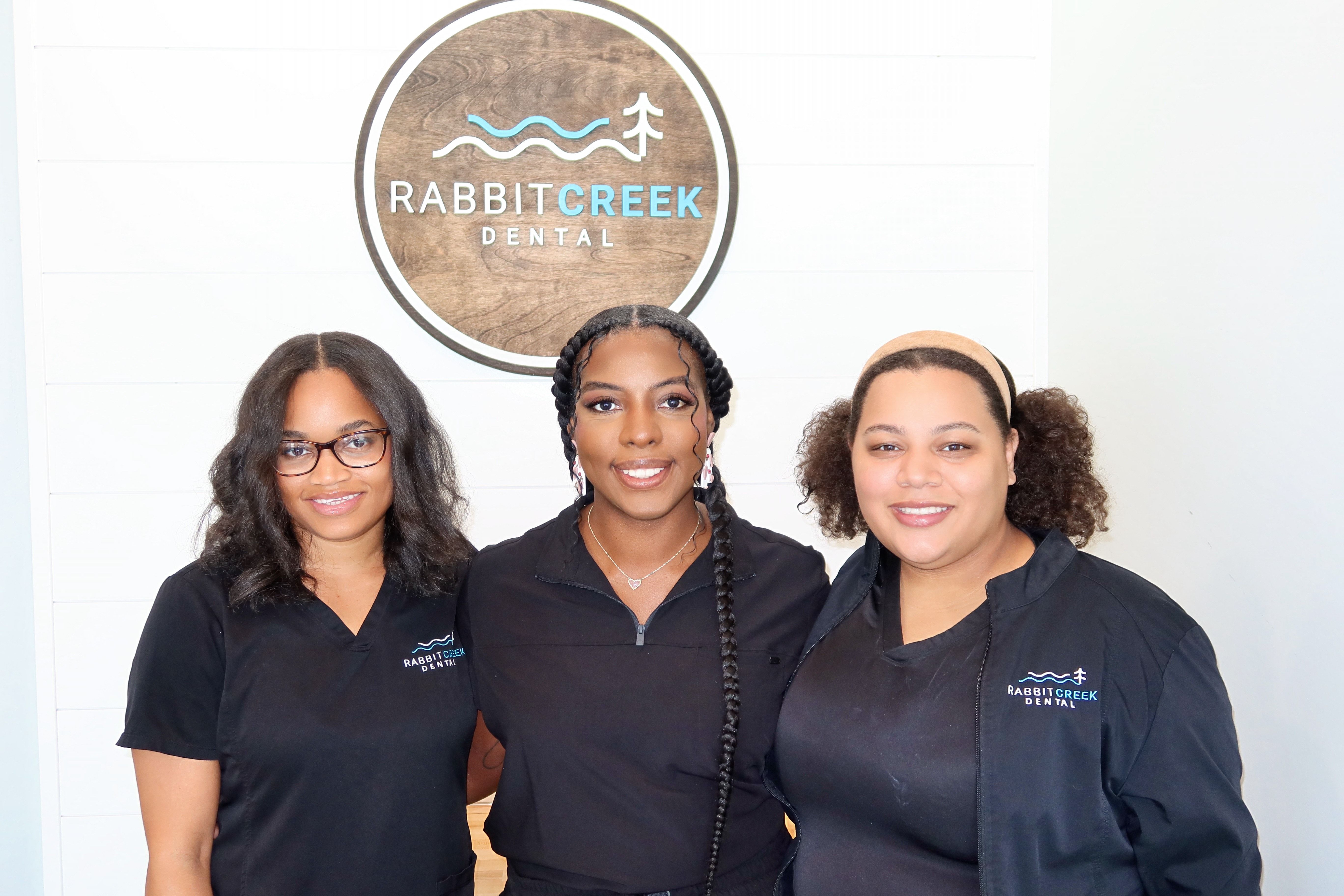 three women are posing for a picture in front of a rabbit creek dental sign .