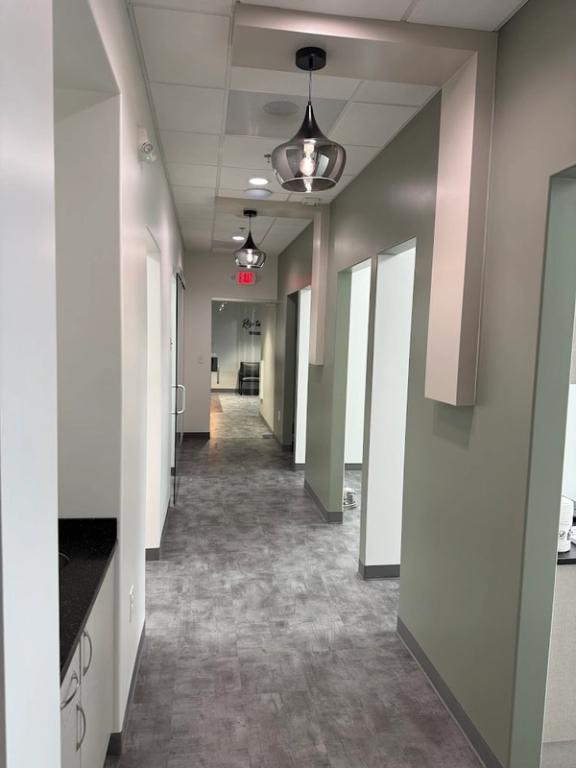 A long, modern hallway with grey floors, white and sage green walls, and three contemporary pendant lights leading to an exit sign in the distance.