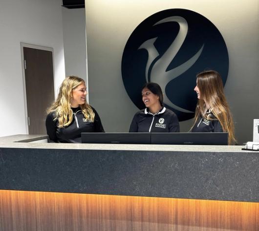 Three smiling women in RISE shirts at a reception desk, with a large RISE logo behind them.