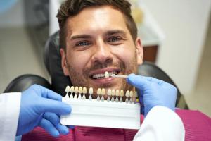 A dentist in blue gloves using a tooth shade guide to match a smiling man's teeth.