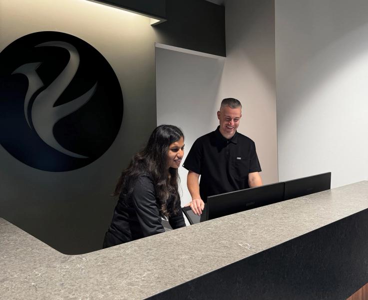 Two smiling staff members look at computer monitors behind a modern reception desk, with a company logo on the wall.