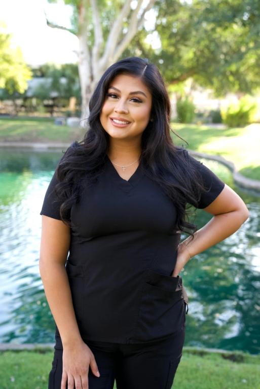 A smiling young woman in black scrubs stands outdoors in front of a pond and trees.