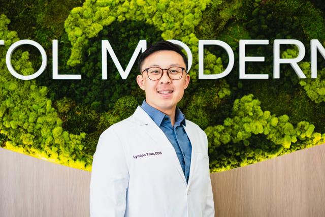 A smiling man, Lyndon Tran, DDS, in a white lab coat stands against a green moss wall with white "MODERN" lettering.