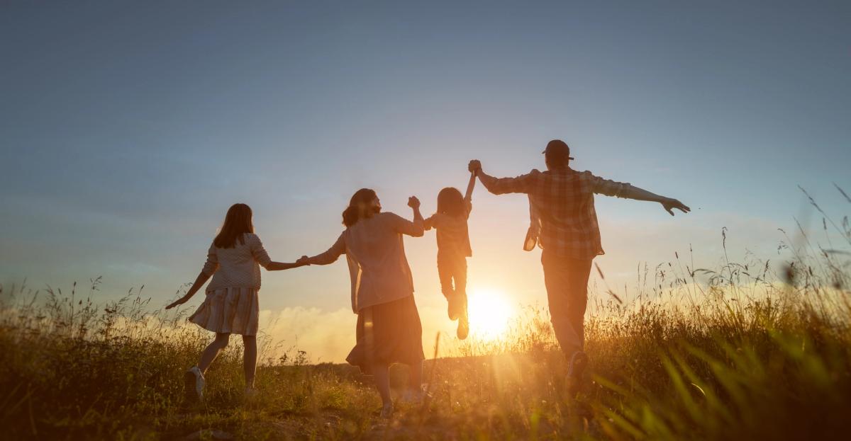 Silhouetted family of four running in a grassy field at sunset, swinging a child.