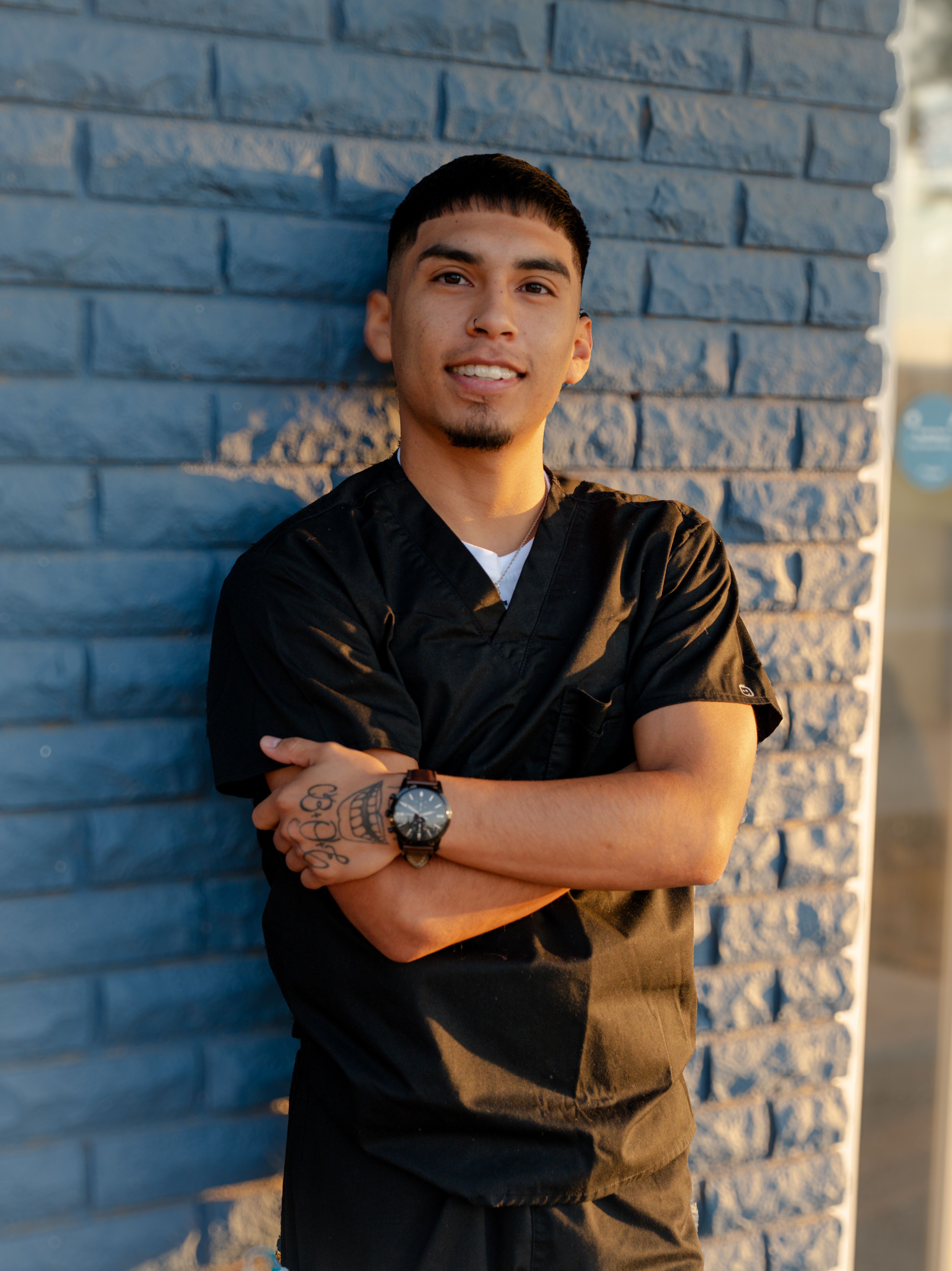 a young man in a black scrub is standing in front of a blue brick wall with his arms crossed .
