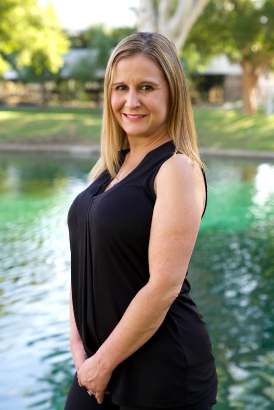 A smiling blonde woman in a black top stands outdoors beside a pond.