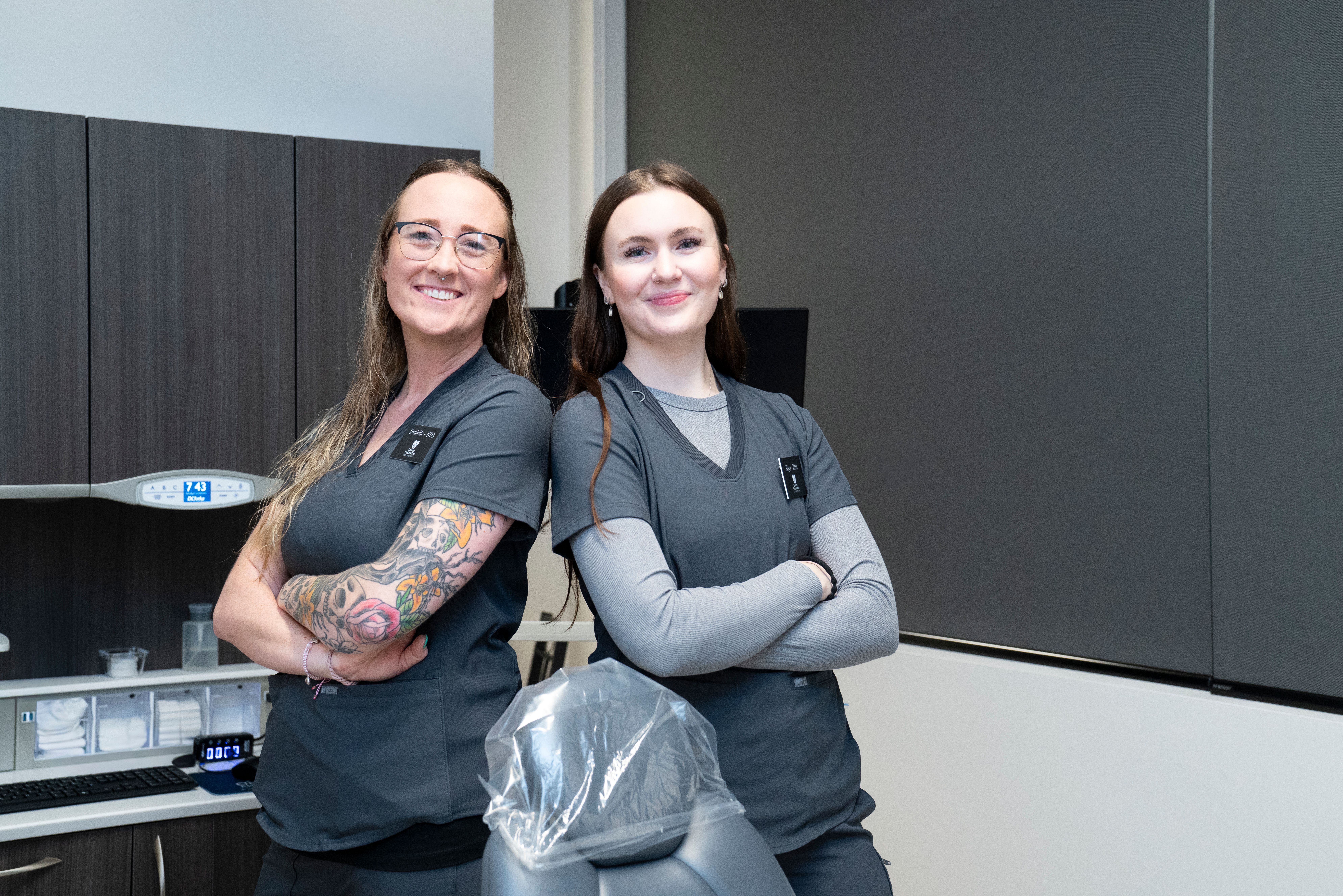 Two smiling women in grey scrubs stand with crossed arms in a modern clinic.