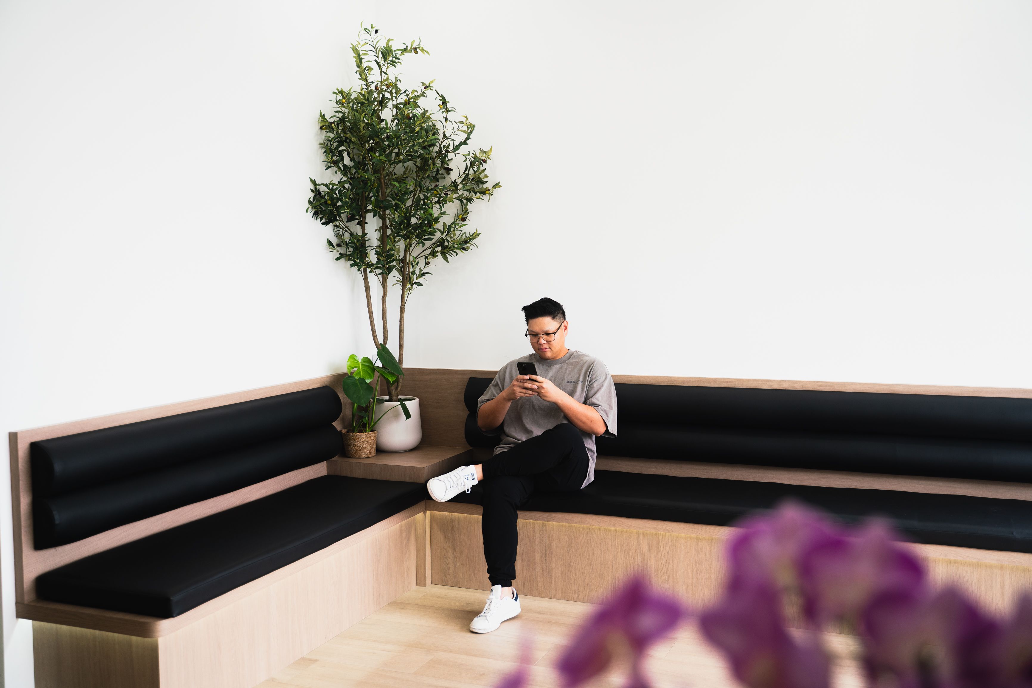 A man sits on a modern L-shaped wooden bench with black cushions, looking at his phone. A large plant is in the corner.