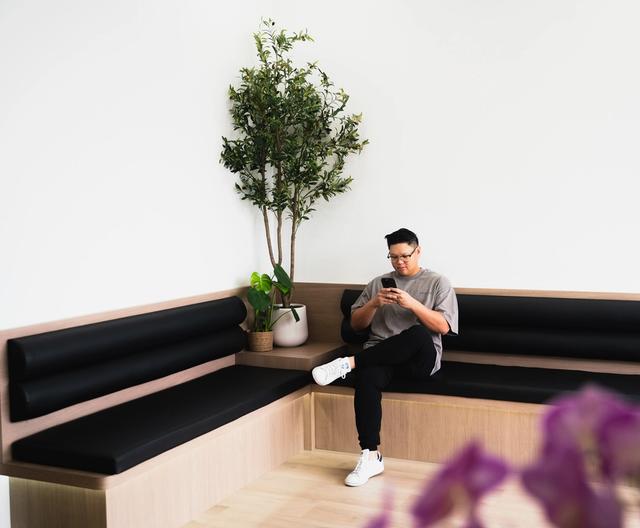 A man sits on a modern L-shaped wooden bench with black cushions, looking at his phone. A large plant is in the corner.