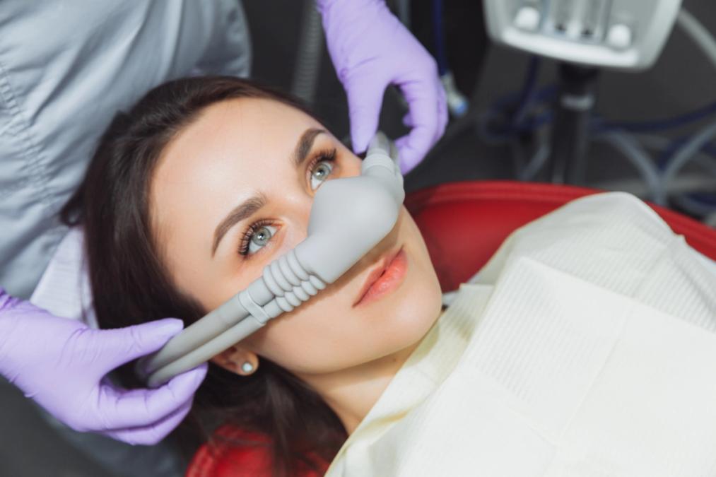 Woman in a dental chair with a nose mask for sedation, being adjusted by gloved hands.