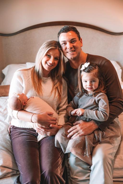 A smiling family of four, with parents holding a swaddled newborn and a toddler, sits together on a bed.