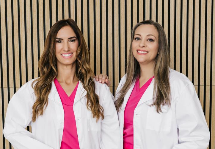 Two smiling women in white lab coats and pink scrubs stand against a striped wooden wall.