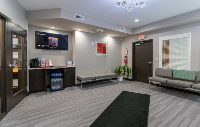 Modern waiting room with a beverage station, TV, gray benches, and patterned carpet.