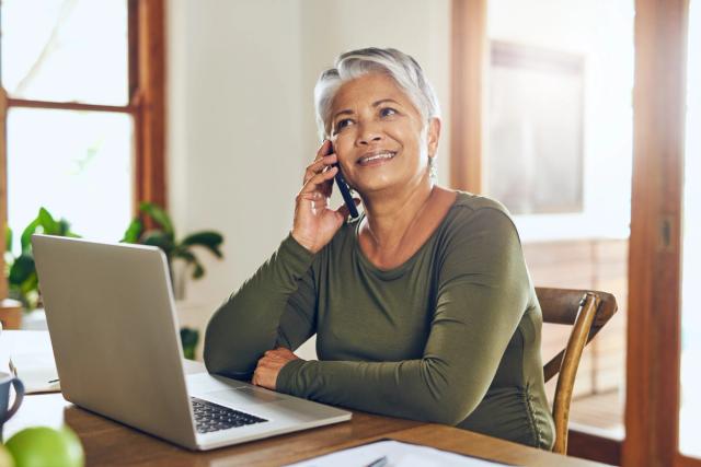 an older woman is sitting at a table talking on a cell phone in front of a laptop computer .