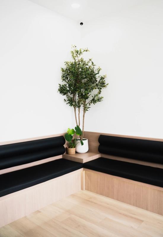 Light wood built-in corner seating with black cushions and potted plants against white walls.