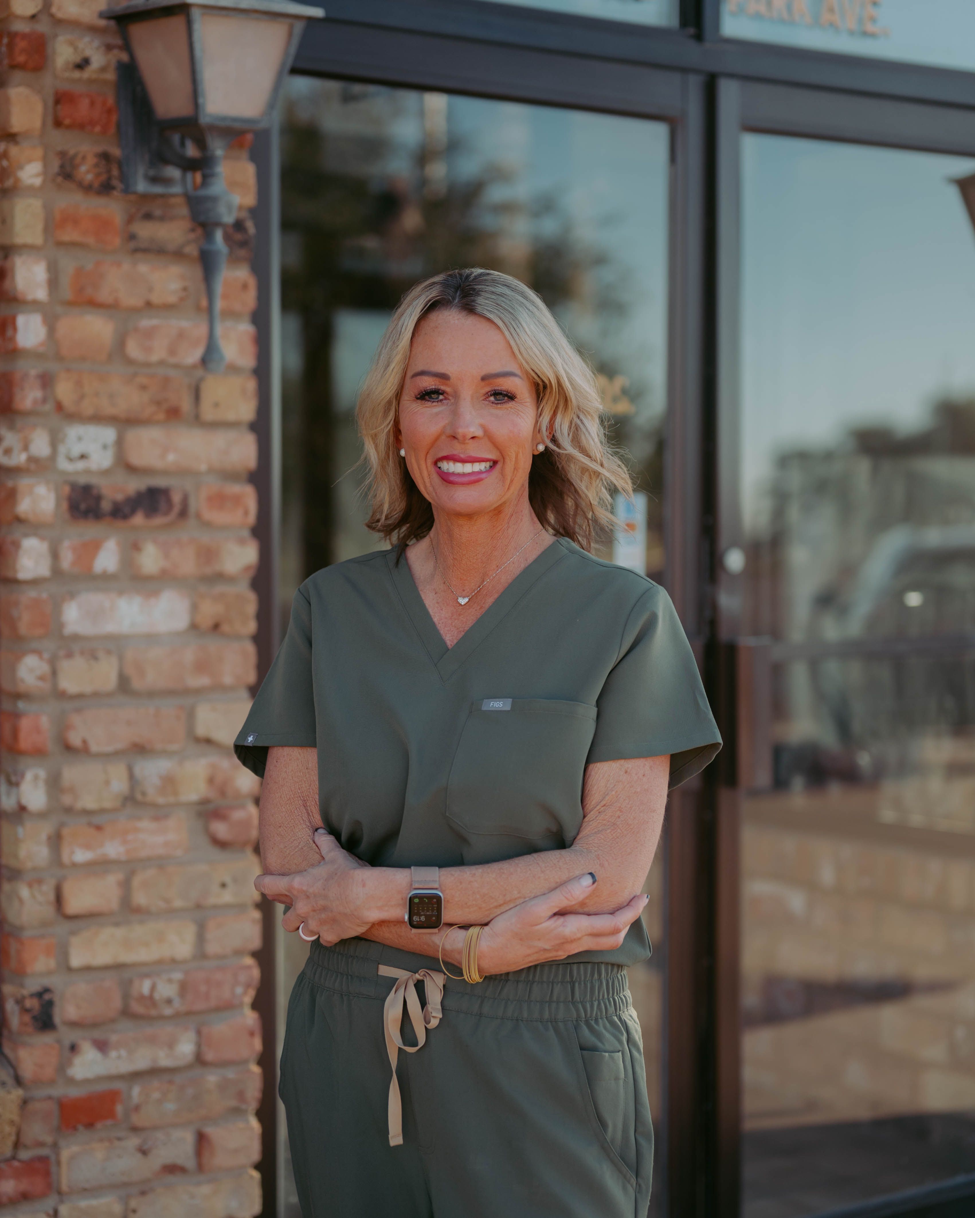 A smiling woman with blonde hair in green scrubs stands with arms crossed in front of a brick building and glass door.