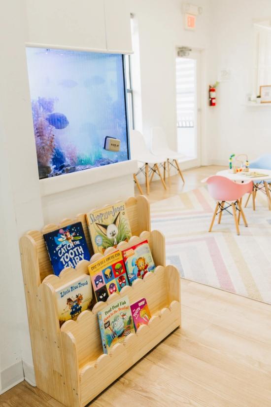 A bright children's play area with a wooden bookshelf filled with books, a large built-in fish tank, and small tables and chairs on a colorful striped rug.