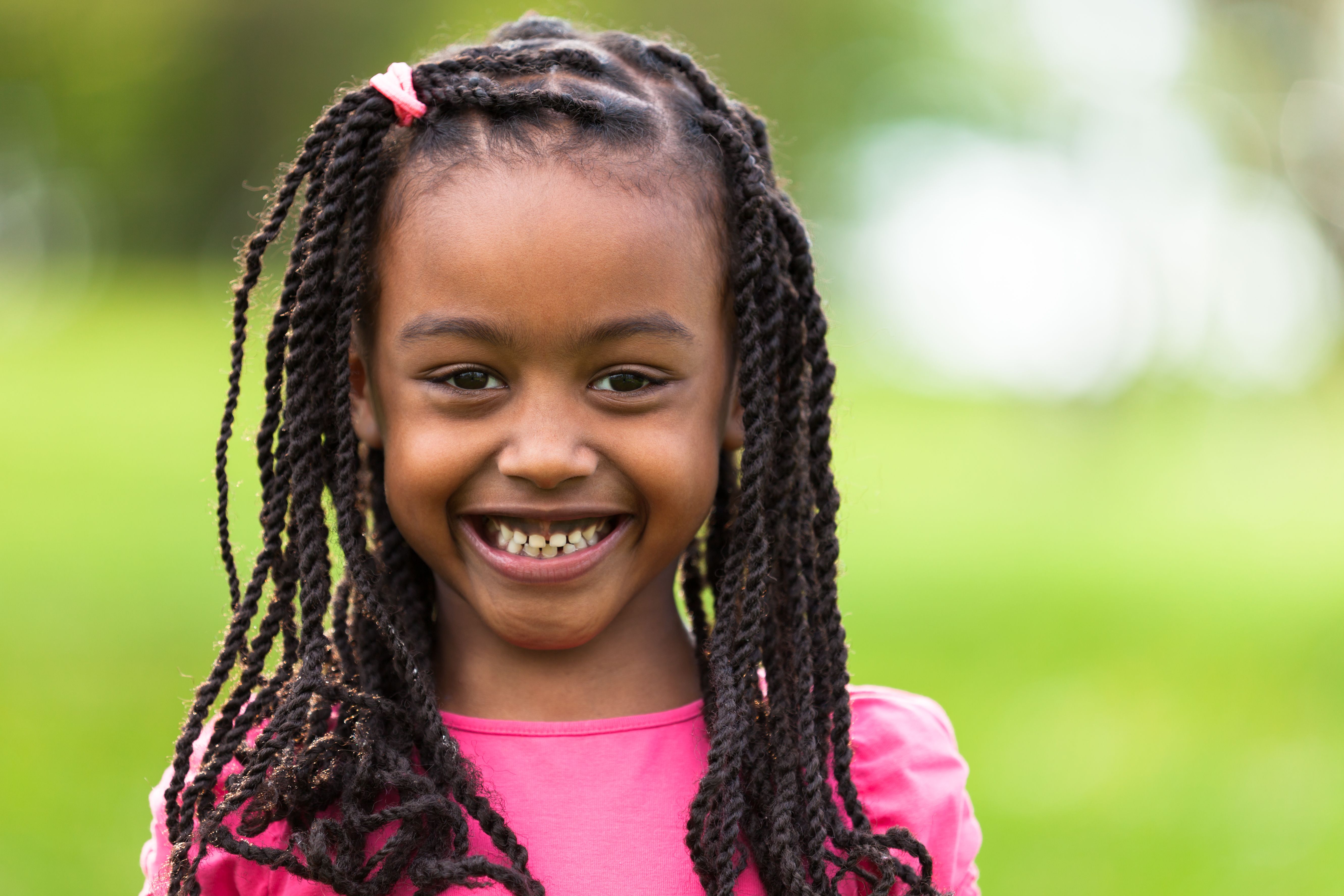A smiling young Black girl with long braids and a pink shirt stands in front of a blurred green background.