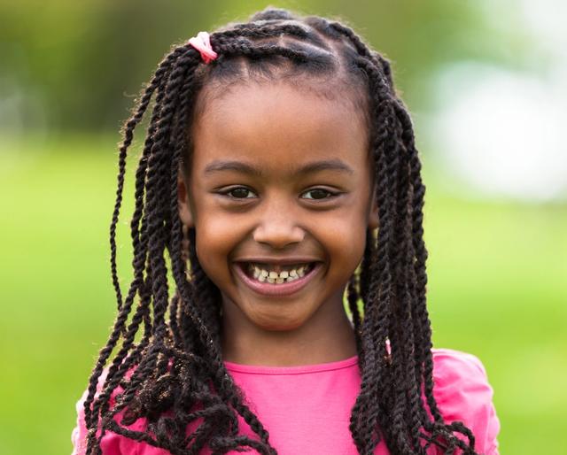 A smiling young Black girl with long braids and a pink shirt stands in front of a blurred green background.