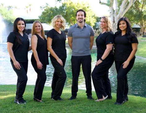 Six smiling team members, five women in black scrubs and one man in a gray polo, standing by a pond with a fountain.