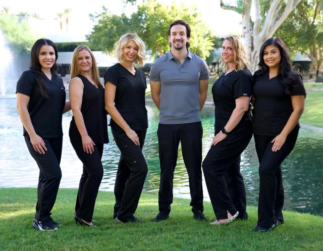 Six smiling people, five women in black scrubs and one man in a grey polo, stand on grass in front of a pond with a fountain.
