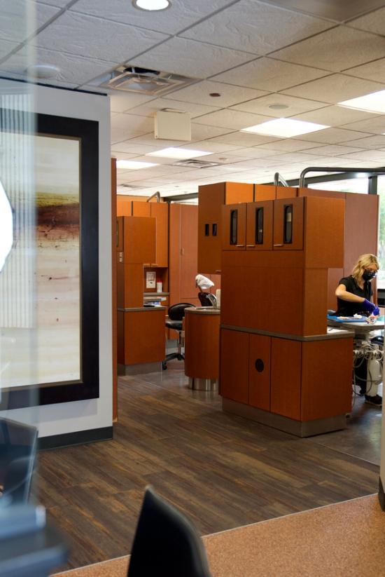 A dental office with several individual treatment bays featuring brown cabinets, where a masked assistant prepares instruments.
