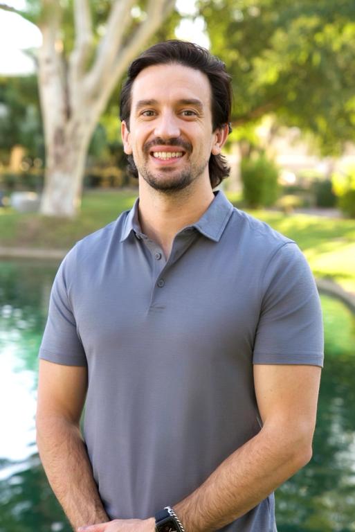 A smiling man in a gray polo shirt stands outdoors with a blurred background of trees and water.