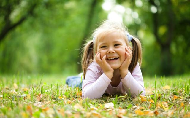 A happy young girl with pigtails lies on green grass, smiling with her chin in her hands.