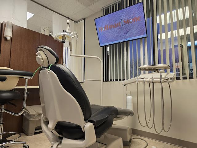 An empty dental examination room with a black and white dental chair, instruments, and a monitor displaying "Hoffman Stone".