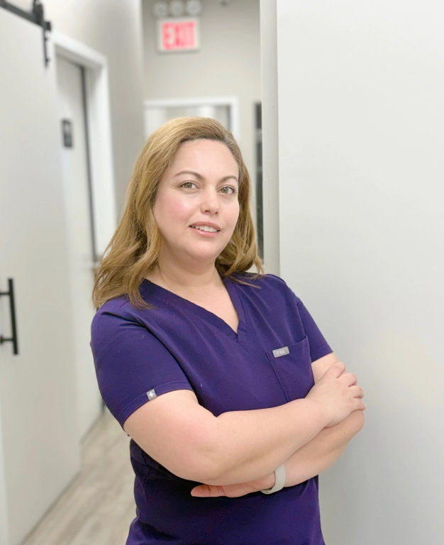 A smiling woman in purple scrubs with crossed arms stands in a hallway.