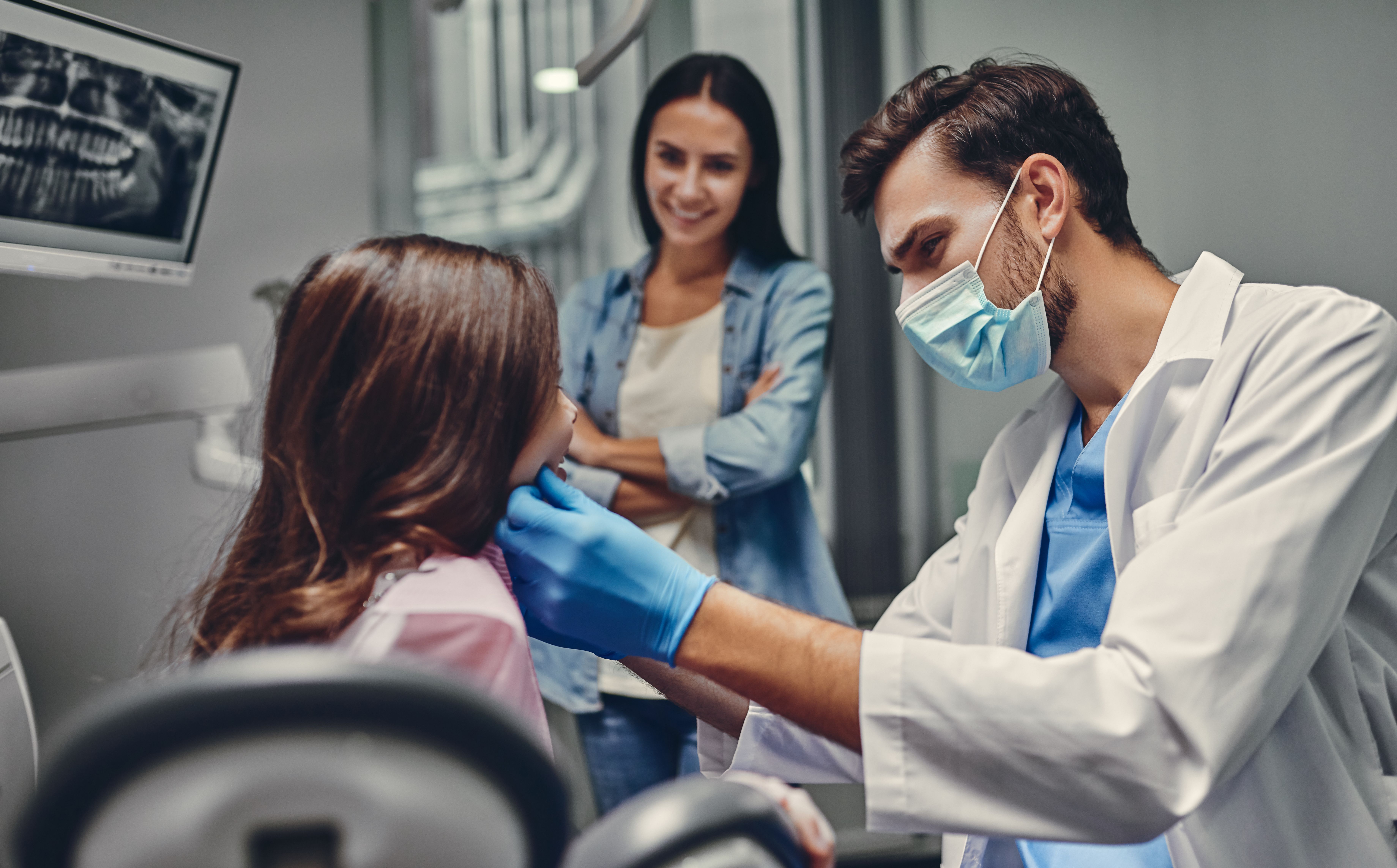 A dentist examines a child's mouth while her mother smiles in the background.