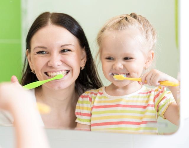 A smiling mother and child reflected in a mirror, both brushing their teeth.