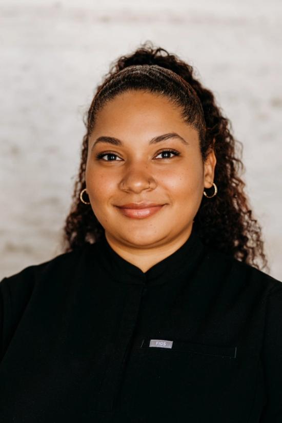 A smiling woman with dark curly hair, wearing a black FIGS uniform top and small gold hoop earrings.