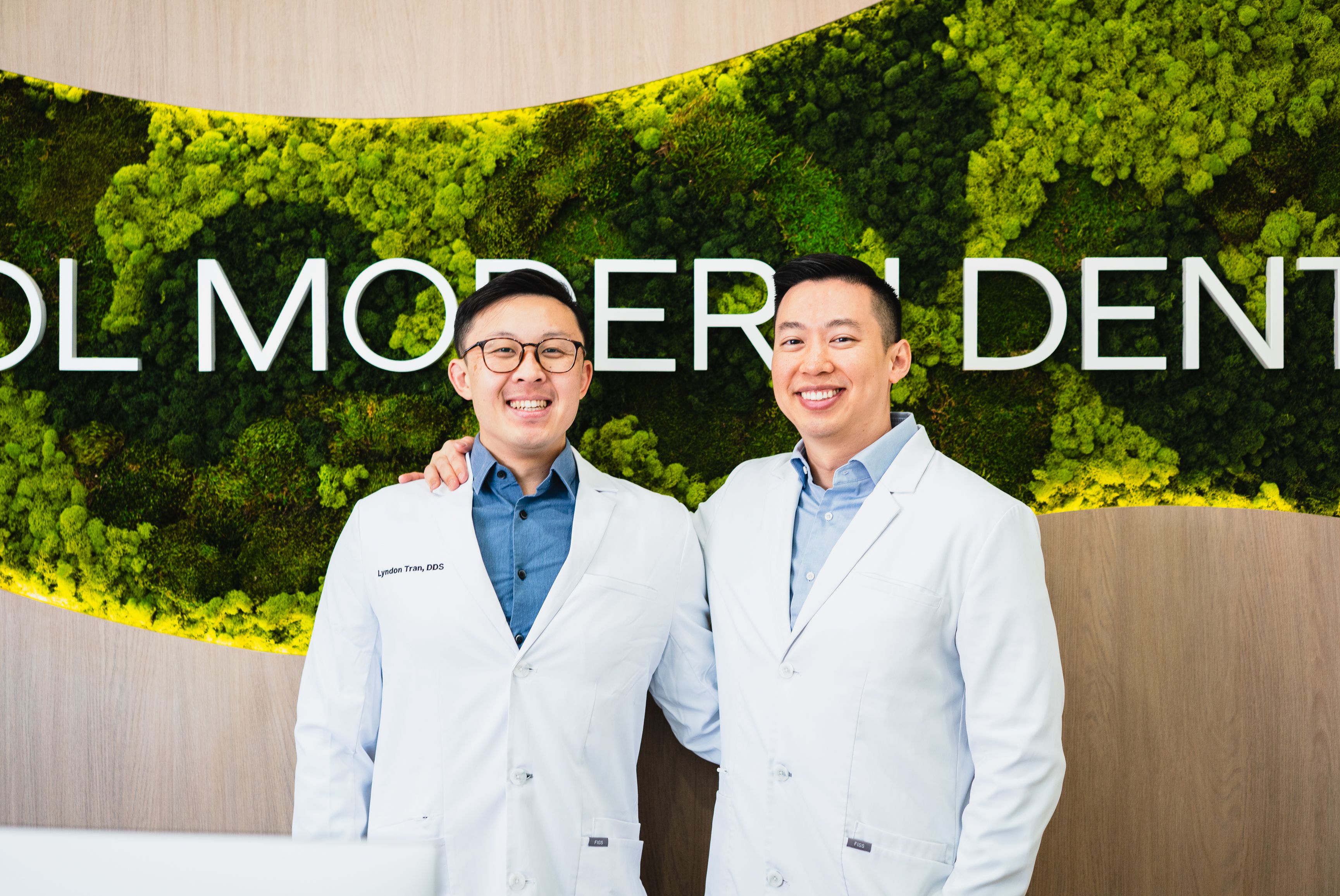 Two smiling dentists in white lab coats, one named Lyndon Tran, standing before a "MODERN DENTAL" sign on a moss wall.