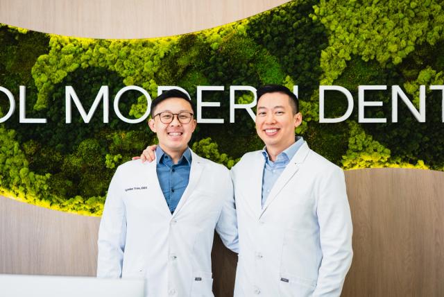 Two smiling dentists in white lab coats stand in front of a green moss wall with a "Modern Dental" sign.
